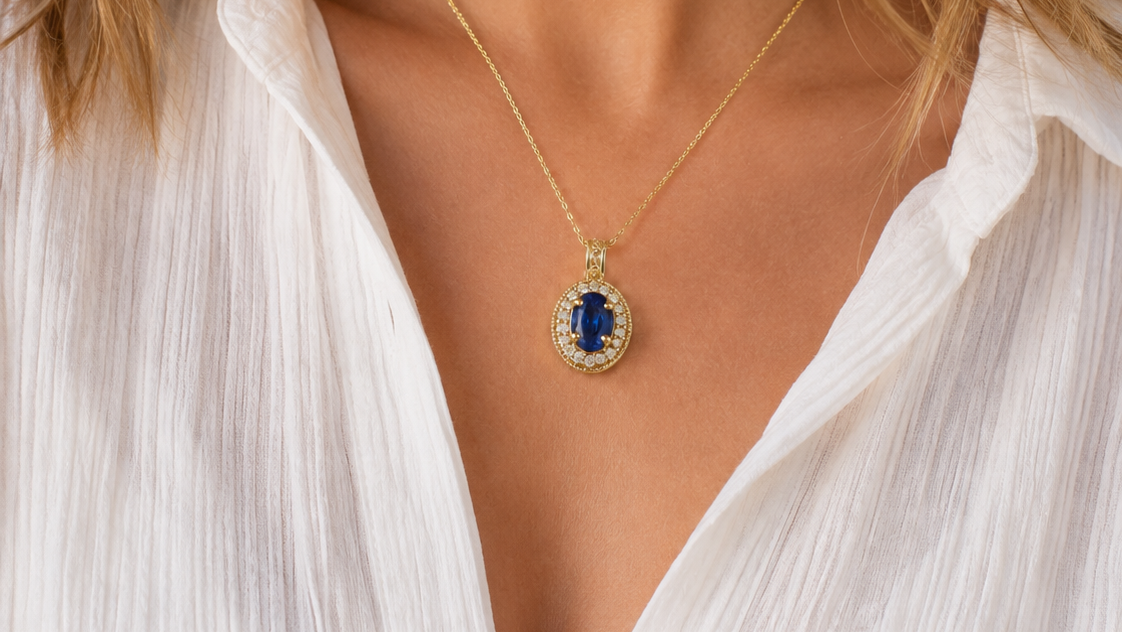 A close-up lifestyle shot of a woman wearing a delicate gold chain necklace with an oval pendant featuring a deep blue center stone surrounded by small clear stones.