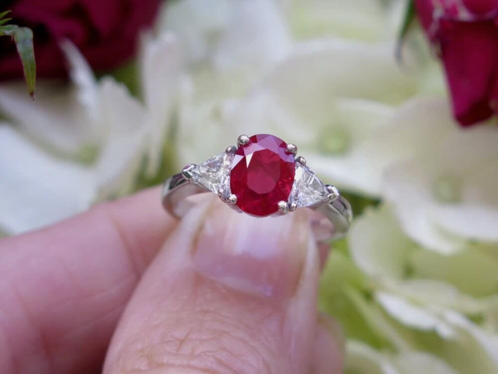 Oval ruby ring with trillion cut sapphires in white gold being held against a somewhat blurred background of white and red roses.