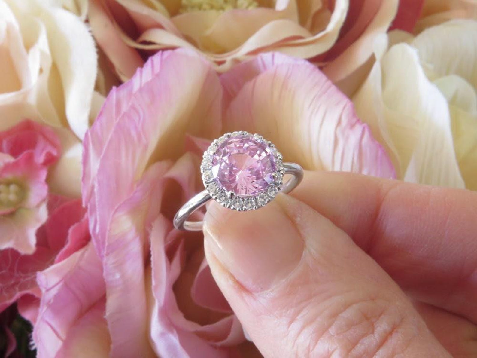 Large, round, light pink sapphire set in delicate diamond halo setting, being held by two fingers in front of a flower.