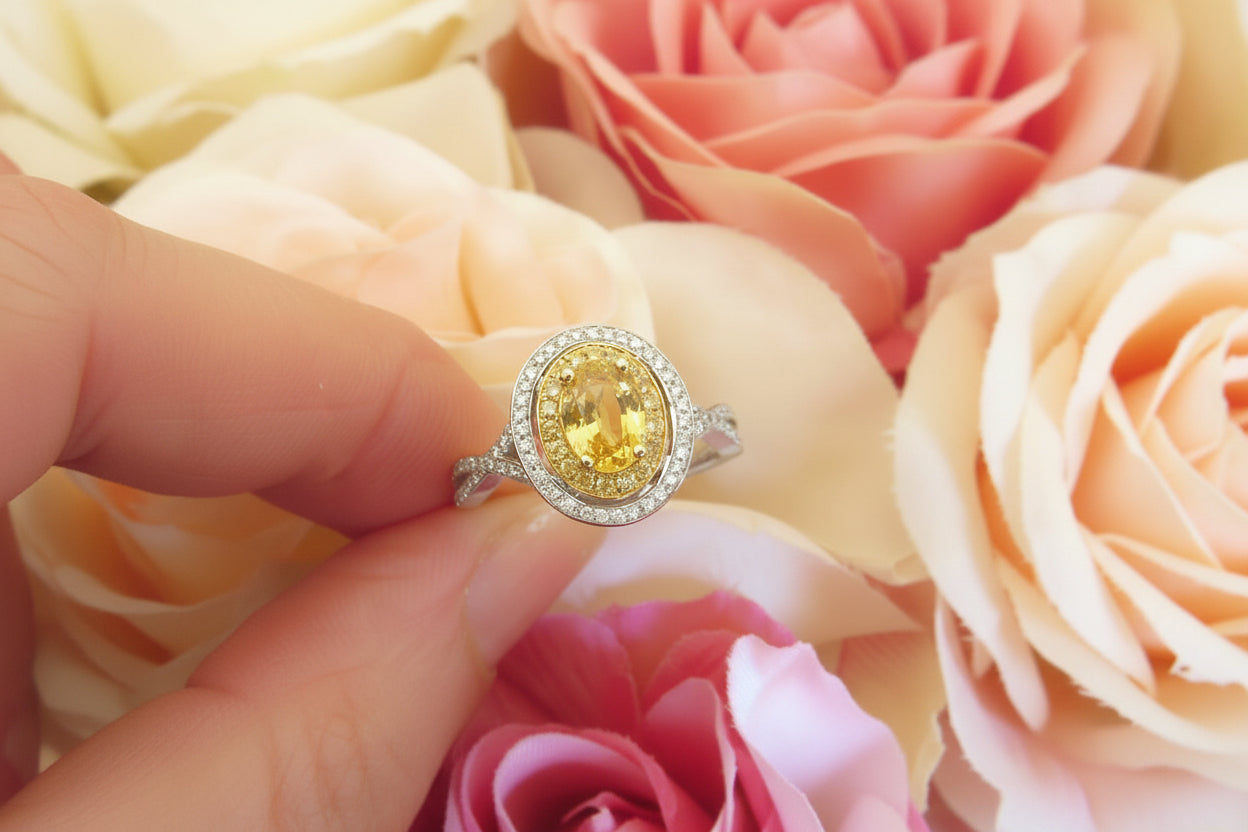A yellow oval ring held between fingers against a soft background of peach and pink roses. The ring features a yellow center stone surrounded by a halo and a silver-colored band with small clear stones.