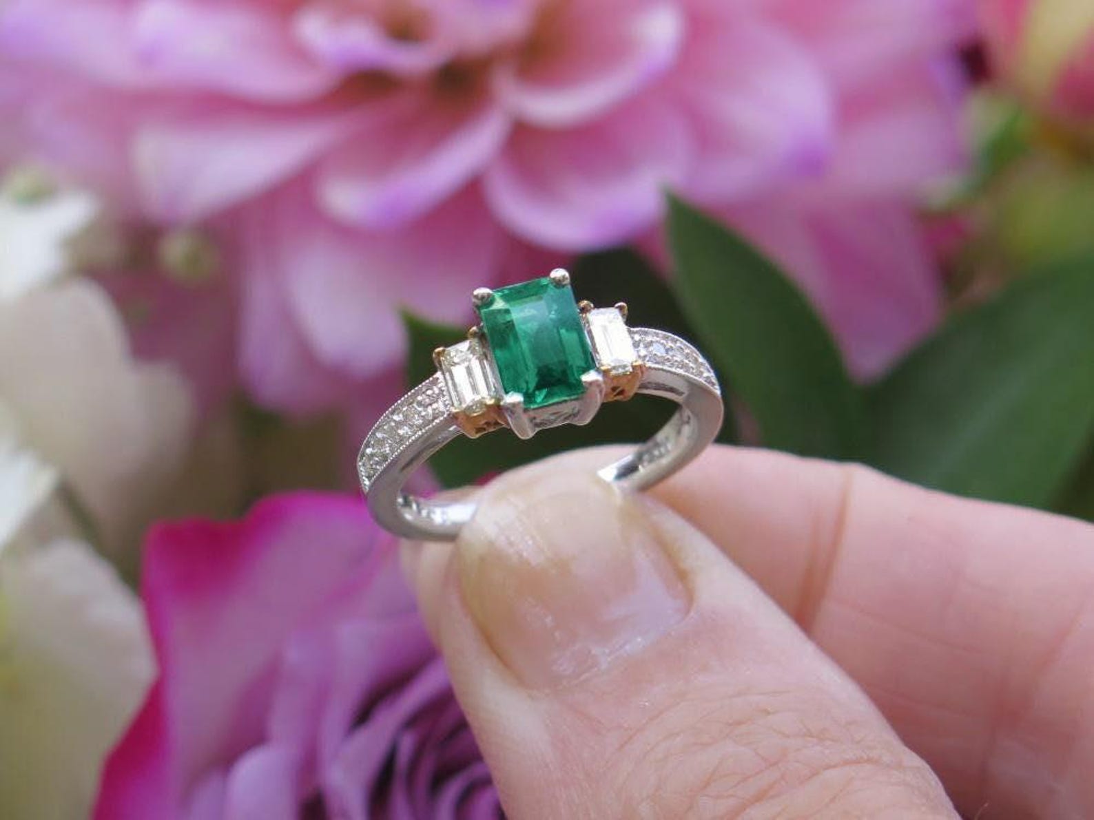 Emerald cut emerald, three-stone engagement ring with emerald cut diamonds, and a diamond-studded band, held against a blurred, colorful flower arrangement.