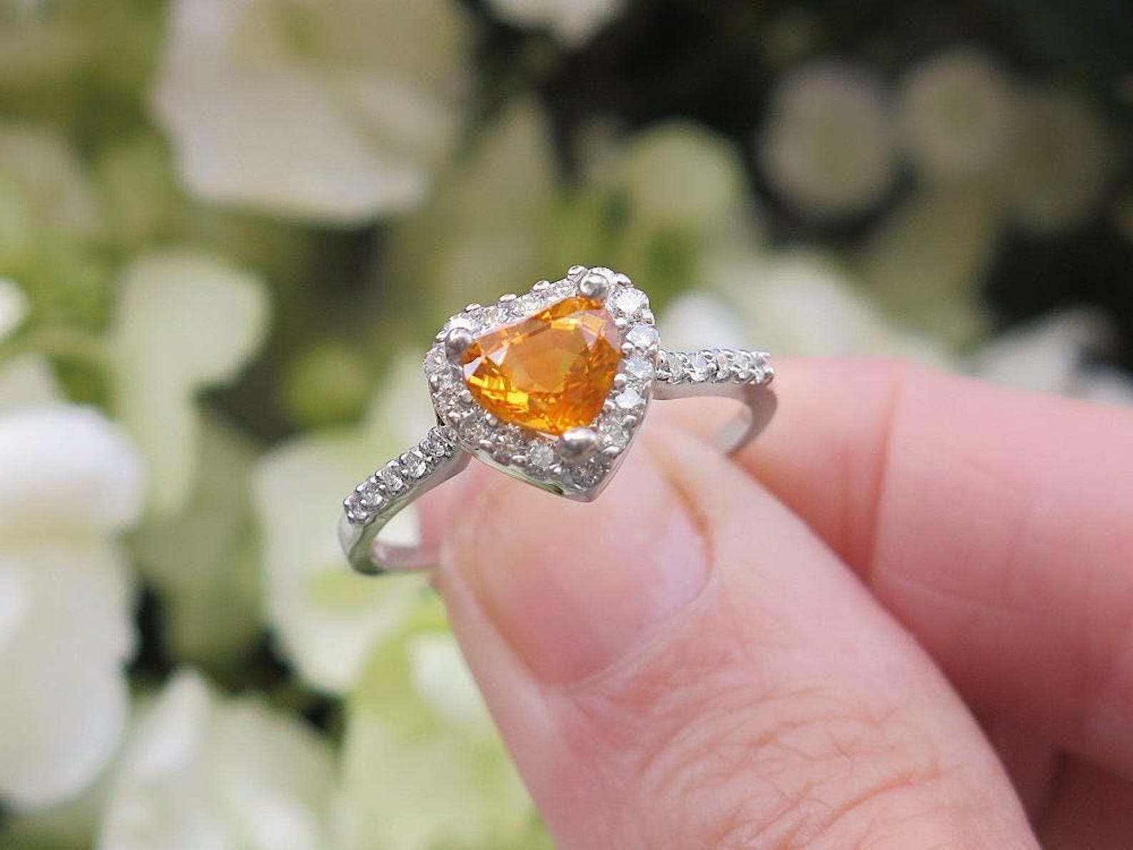 Heart cut, orange sapphire engagement ring with diamond halo, against a blurred white flower background.