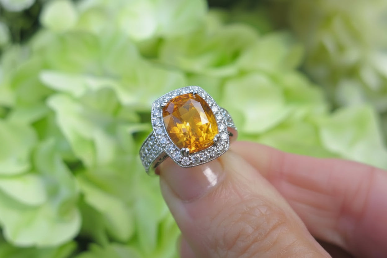 A white gold ring with a large oval orange sapphire gemstone and a halo of small clear diamond stones, held between fingers against a soft-focus background of light green hydrangea flowers.