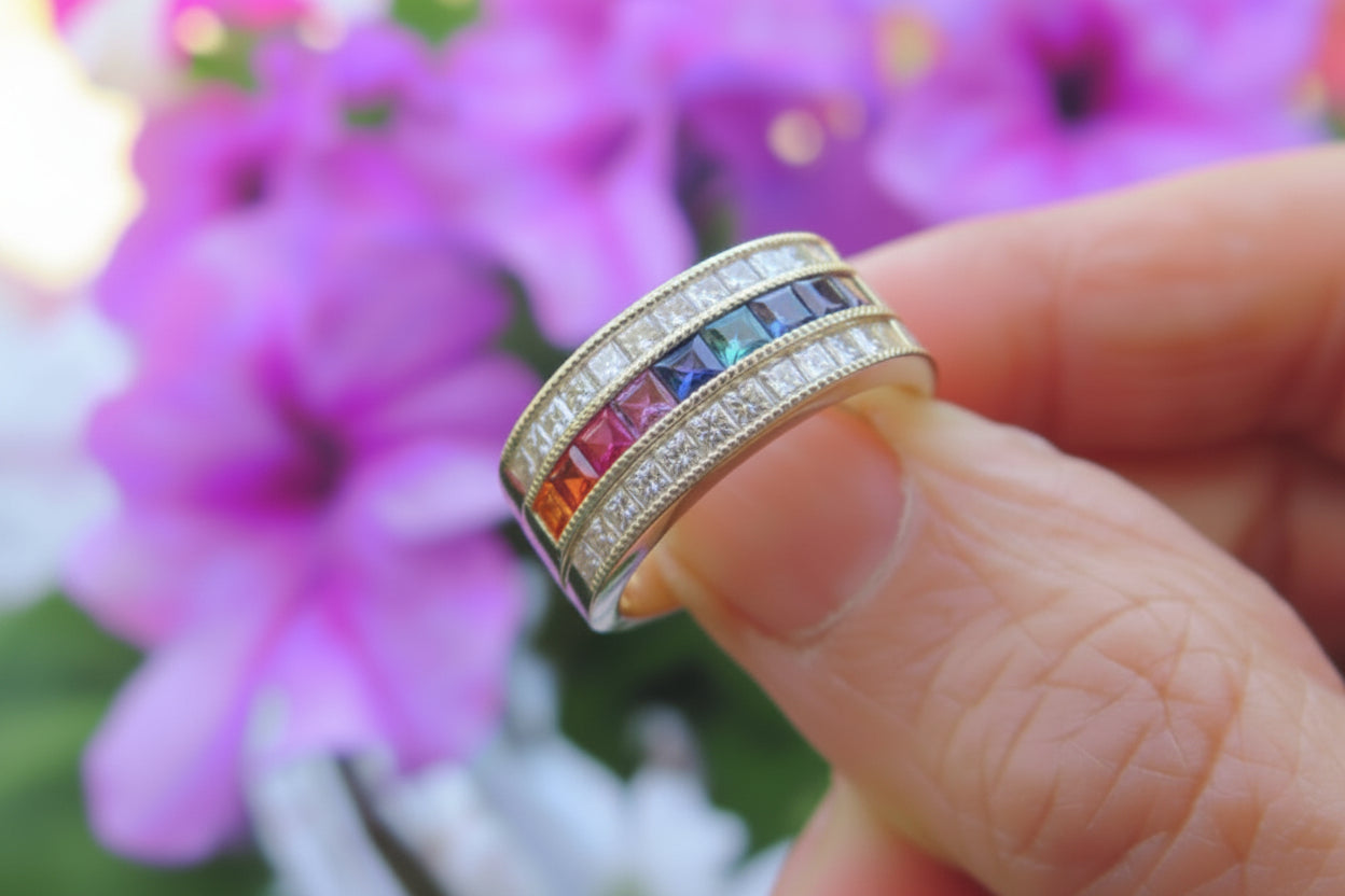 A close-up of a hand holding a yellow gold ring with three rows of colorful sapphires, including orange, pink, red, blue, and clear stones, set against a blurred background of pink flowers.