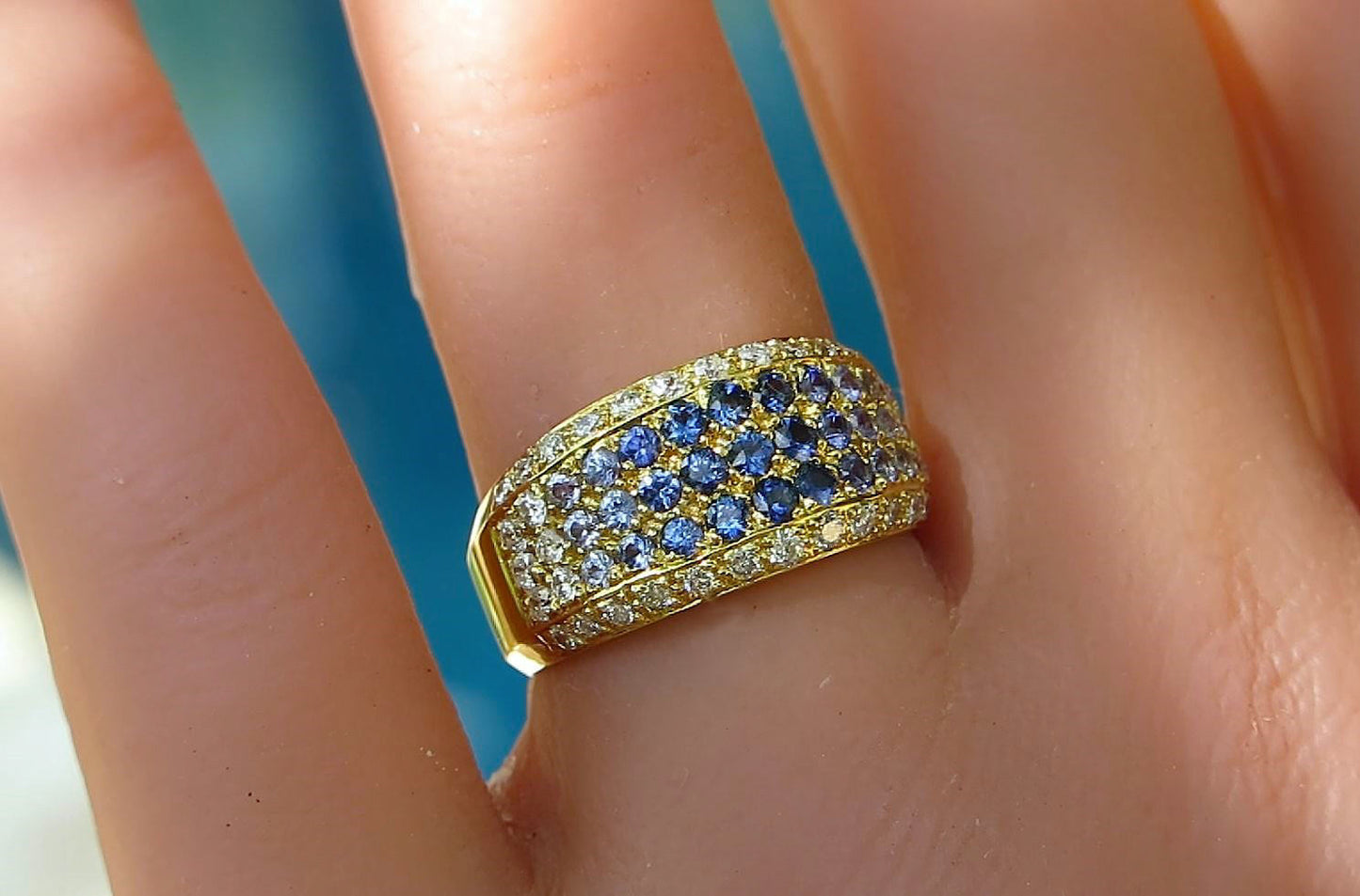 A close-up of a gold band ring with rows of blue and clear pavé stones, worn on a person's finger against a blurred blue background.