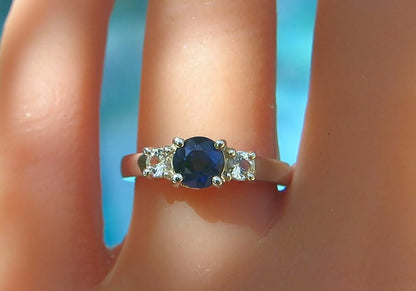 A three-stone ring with a round dark blue center stone and two small clear side stones, shown worn on a finger against a bright blue background.