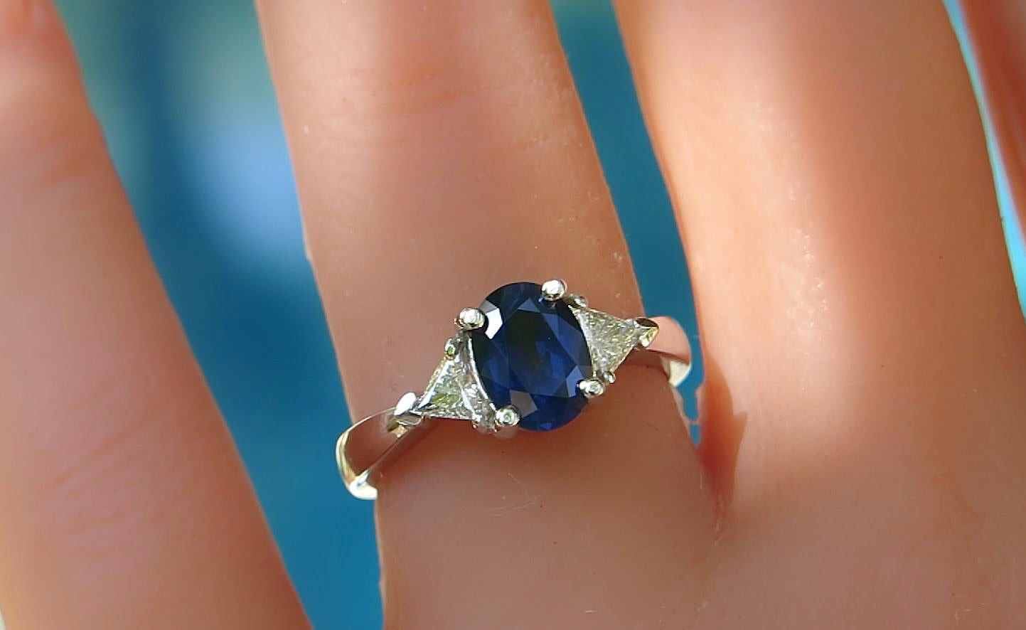 A white gold three stone ring with an oval blue gemstone and small clear accent stones, shown worn on a finger against a blurred blue background.