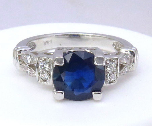 White gold ring featuring a large round-cut deep blue sapphire center stone, accented by small clear diamonds on either side of the band, shown on a plain white background.