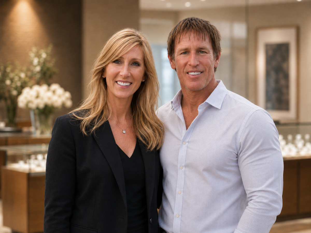 A smiling blonde woman in a black blazer and a smiling man in a light blue button-down shirt standing together in a softly lit jewelry showroom.
