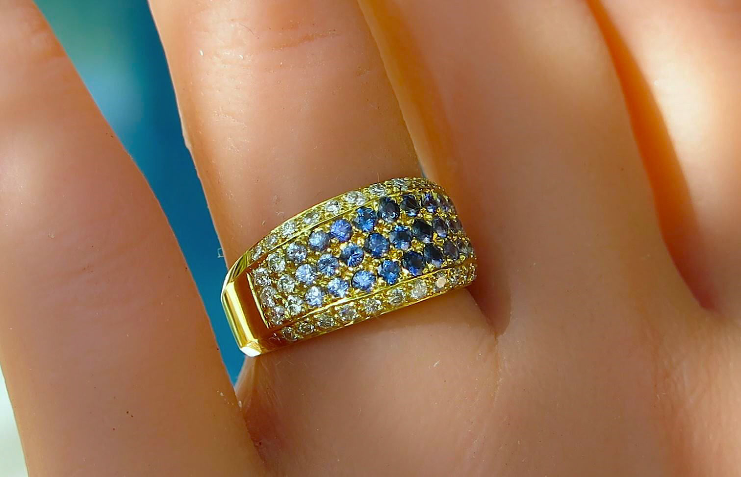 A close-up of a gold band ring with rows of blue and clear pavé sapphires and diamonds, worn on a finger.