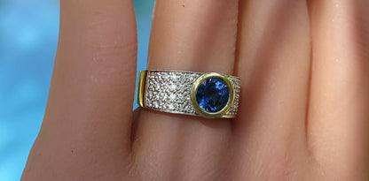 A close-up of a ring worn on a finger, featuring a round blue gemstone in a gold bezel setting, flanked by pavé-set clear stones on a yellow-gold band.