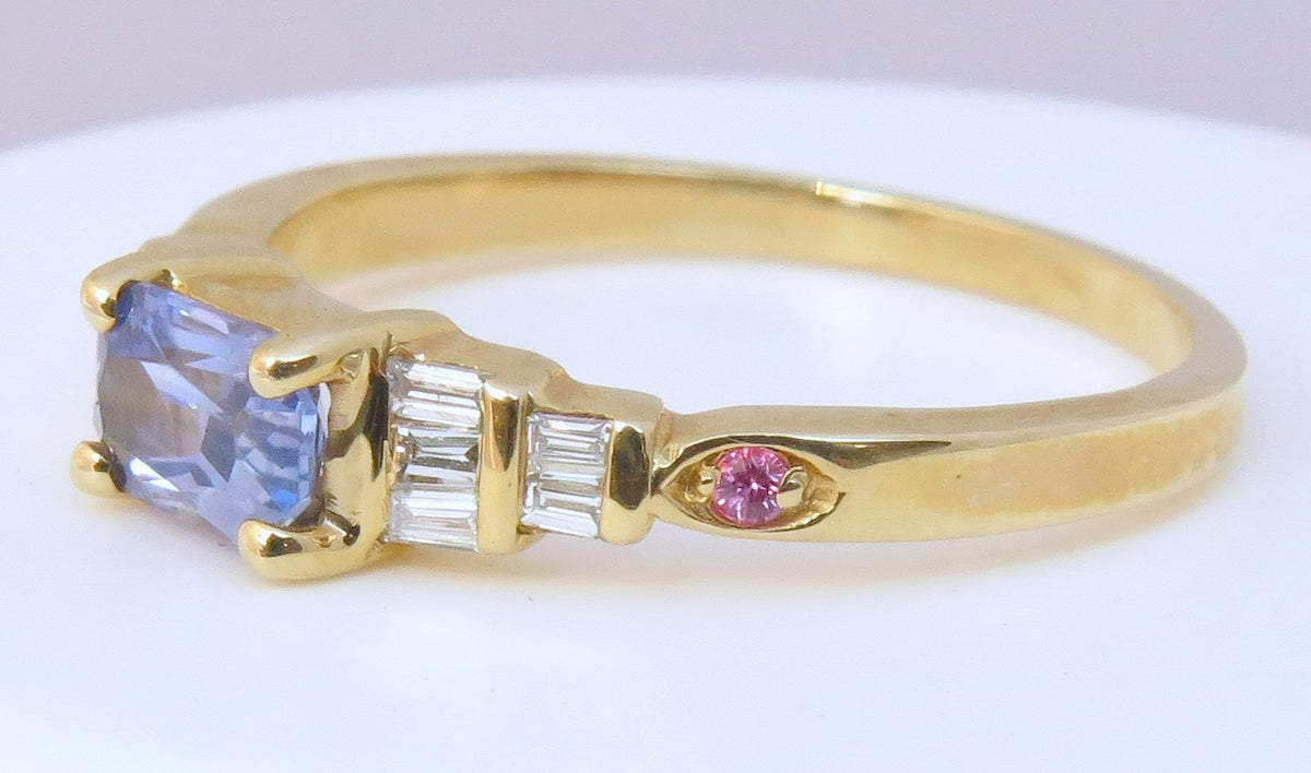 A yellow gold ring featuring a rectangular blue ceylon sapphire center stone, baguette-cut side diamonds, and a small round pink accent stone, shown on a plain white background.