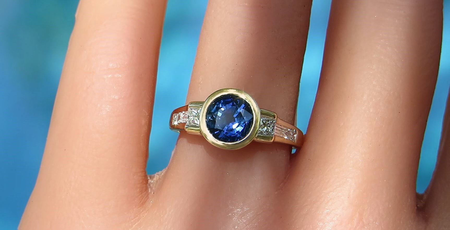A gold ring with a round blue gemstone and small clear diamonds, shown worn on a finger against a bright blue background.
