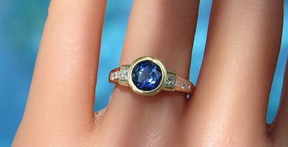 A gold ring with a round blue gemstone and small clear diamonds, shown worn on a finger against a bright blue background.