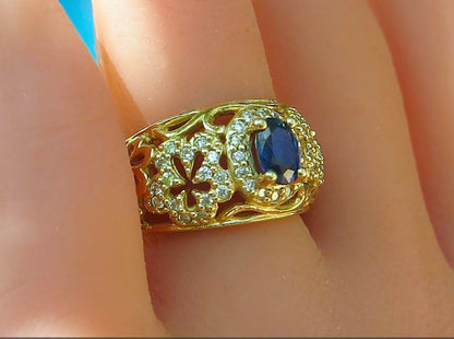 A bold gold floral design ring with a blue oval center stone and small clear stones, shown worn on a finger.