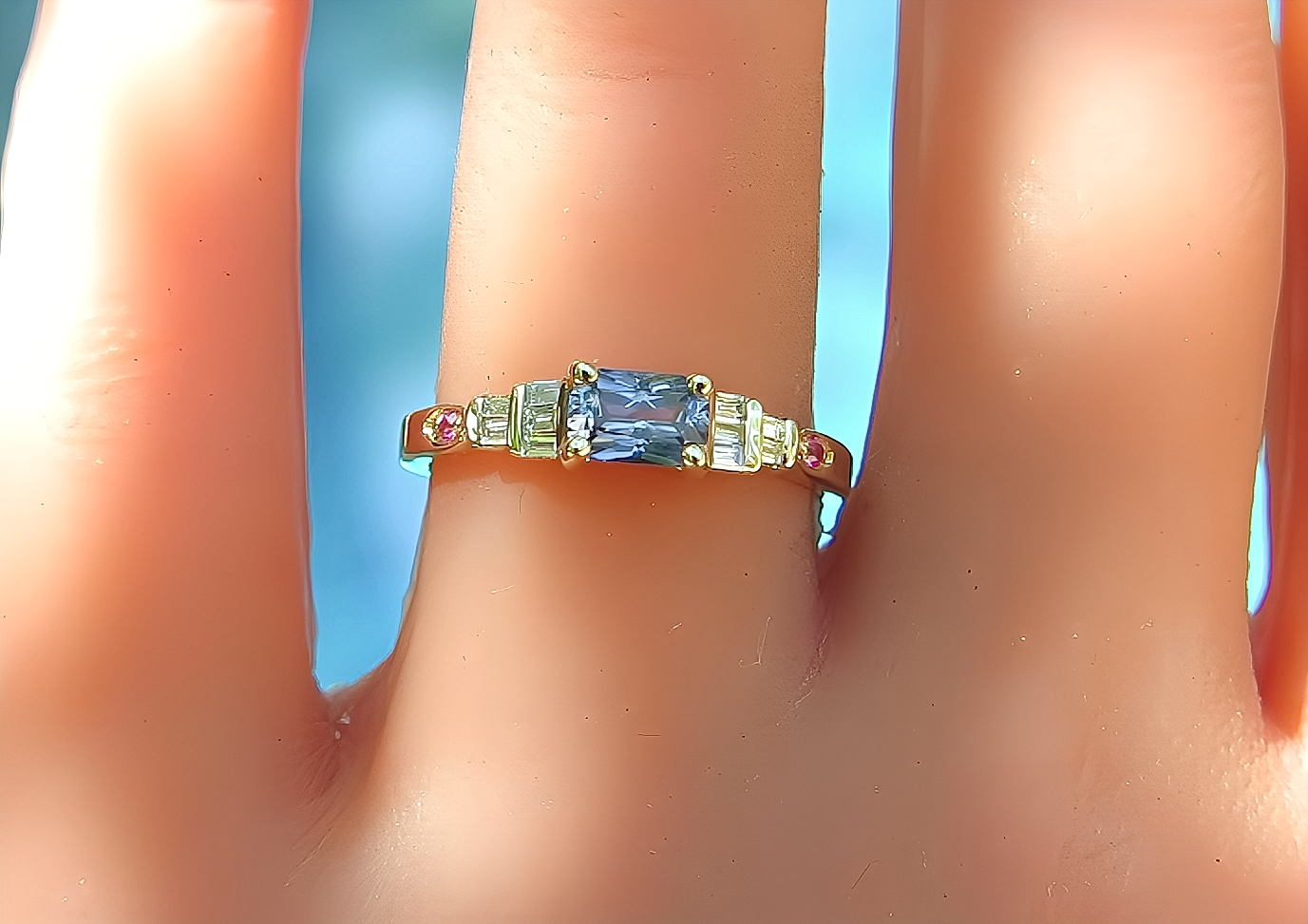 A close-up of a gold ring with a rectangular blue sapphire center stone and baguette-cut side stones, worn on a finger against a bright blue background.