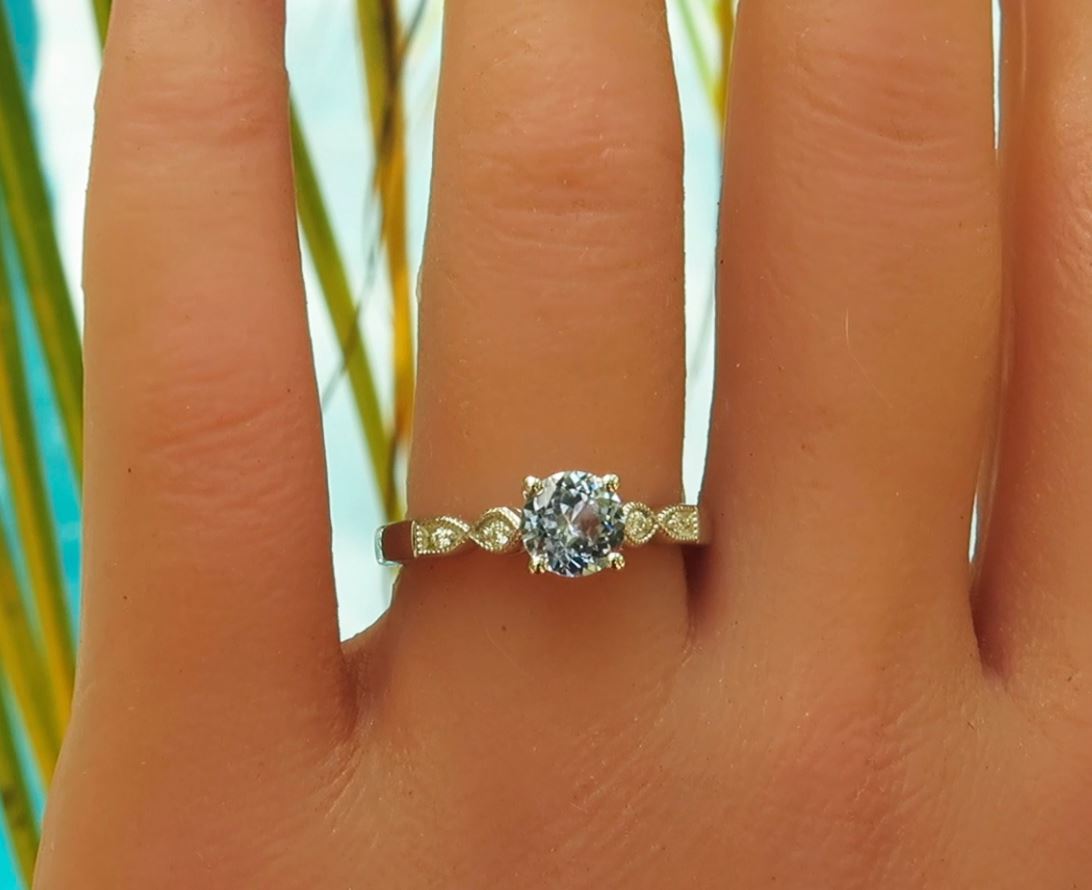 A close-up of a hand wearing a white gold solitaire engagement ring with a round brilliant-cut center stone and small accent diamonds on the band, set against a blurred background of green leaves and light blue sky.