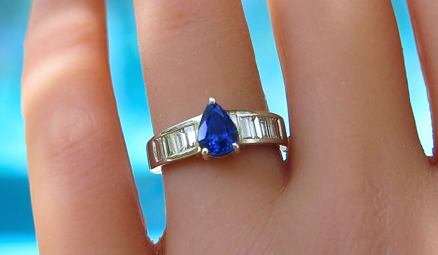 A white gold ring with a pear-cut blue sapphire and baguette side stones, shown worn on a finger against a bright blue background.