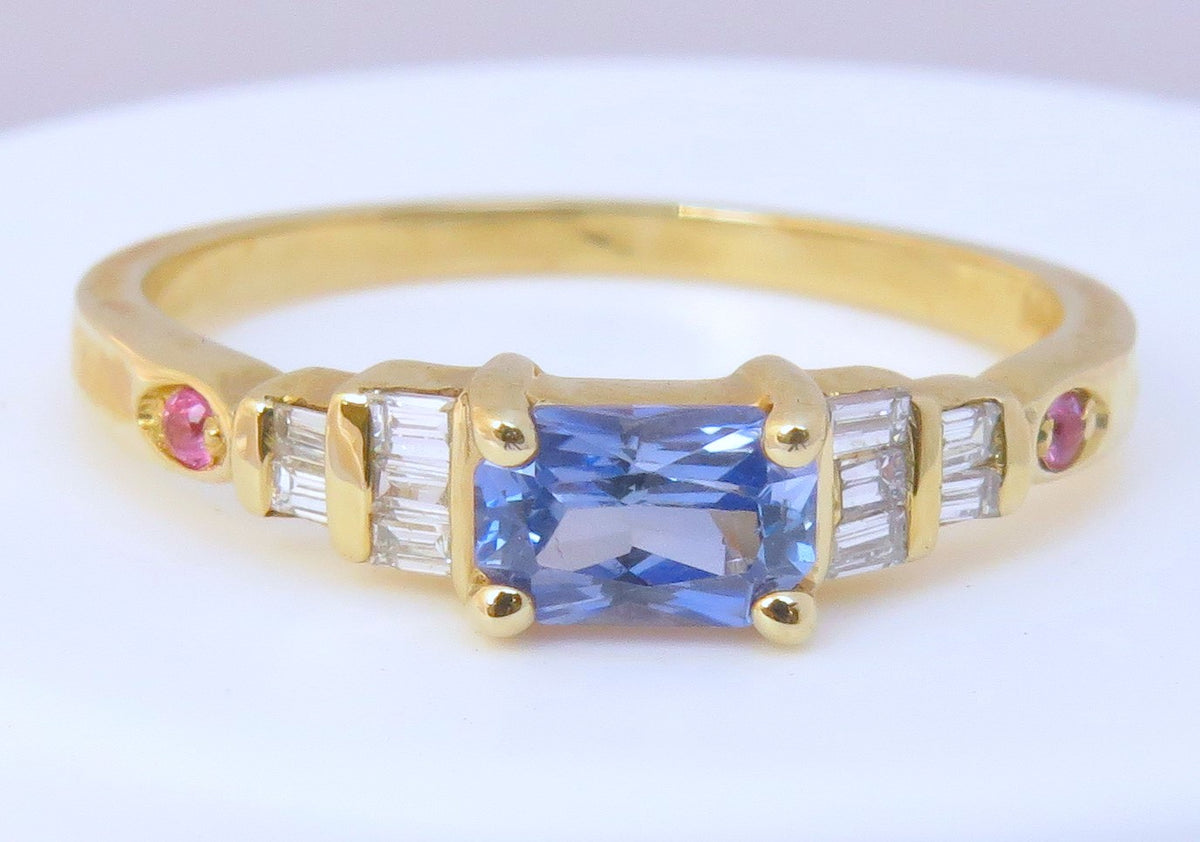 A yellow gold ring featuring a rectangular blue sapphire center stone, flanked by baguette-cut clear diamonds and small round pink gemstones on a white background.