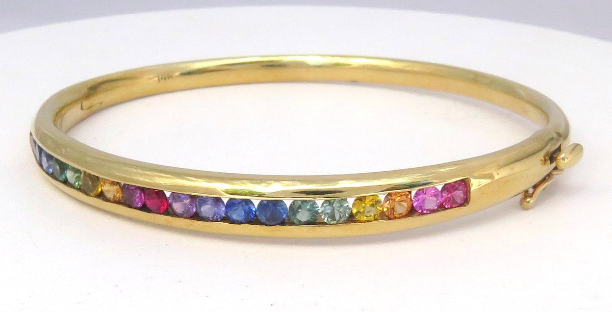 A gold bangle bracelet featuring a row of small, colorful rainbow sapphires channel set , displayed on a plain white background.