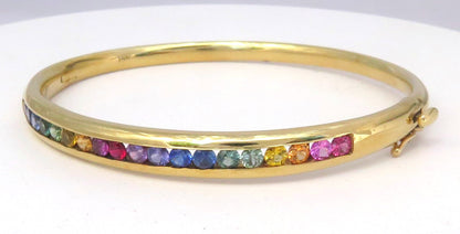 A gold bangle bracelet featuring a row of small, colorful rainbow sapphires channel set , displayed on a plain white background.