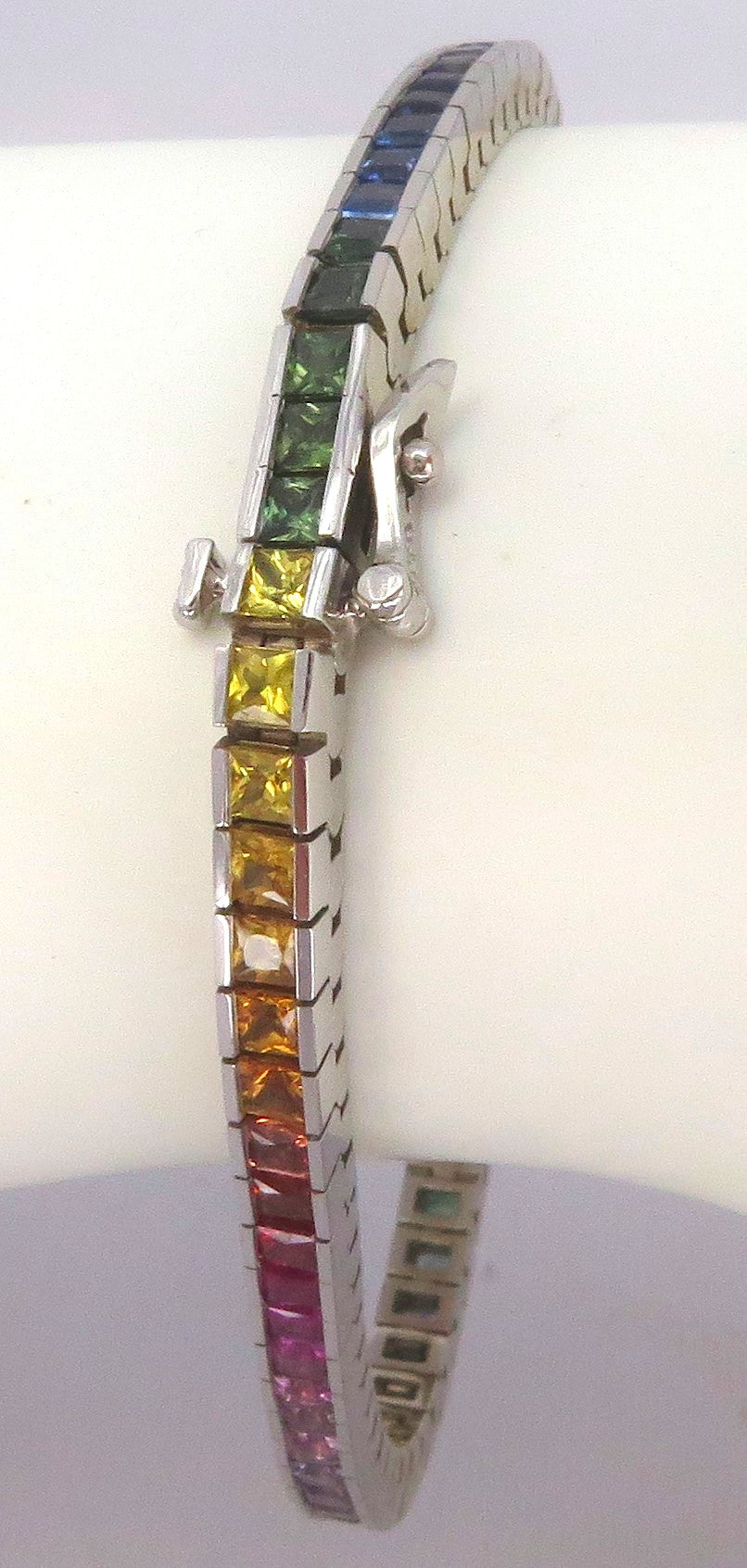 A white gold tennis bracelet with a row of princess square-cut rainbow-colored sapphires, displayed on a white cylindrical stand.