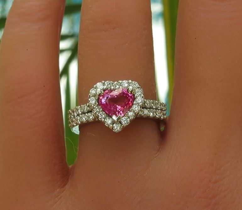 Bright pink sapphire in a heart shape surrounded by diamonds with a diamond encrusted band in platinum on the finger.