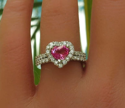 Bright pink sapphire in a heart shape surrounded by diamonds with a diamond encrusted band in platinum on the finger.