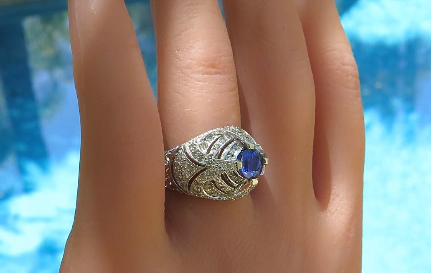 A white gold ring with a central blue gemstone and intricate filigree detailing, shown worn on a person's hand with a blurred blue background.