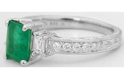 A white gold diamond ring featuring a rectangular green emerald-cut emerald center stone and a row of round diamonds along the band, set against a plain white background.