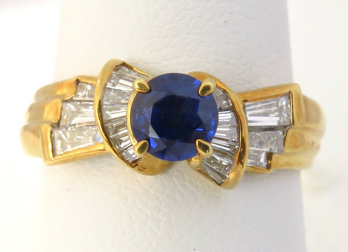 A yellow gold ring featuring a round-cut deep blue sapphire center stone, flanked by baguette-cut clear stones in a fan-like setting, displayed on a white ring stand.