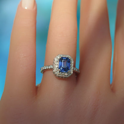 A white gold ring with a rectangular blue sapphire center stone and a halo of small clear stones, shown worn on a finger against a bright blue background.