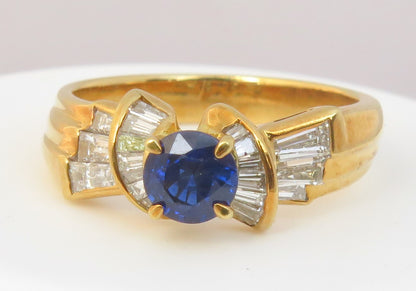A yellow gold ring featuring a round-cut deep blue sapphire center stone, flanked by baguette-cut clear stones in a curved setting, photographed on a plain light background.