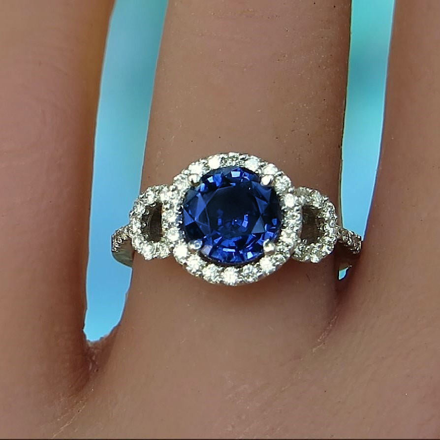 A close-up of a ring worn on a finger, featuring a round-cut deep blue center stone surrounded by a halo of small clear stones, with additional clear stones on the band.