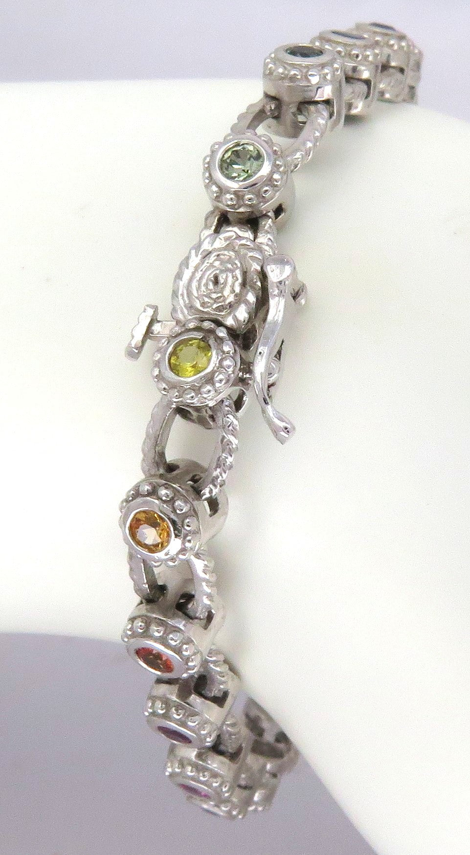 A white gold link bracelet displayed on a white mannequin arm. The bracelet features multiple oval links, alternating with bezel set sapphires in shades of  green, yellow, orange, and red.