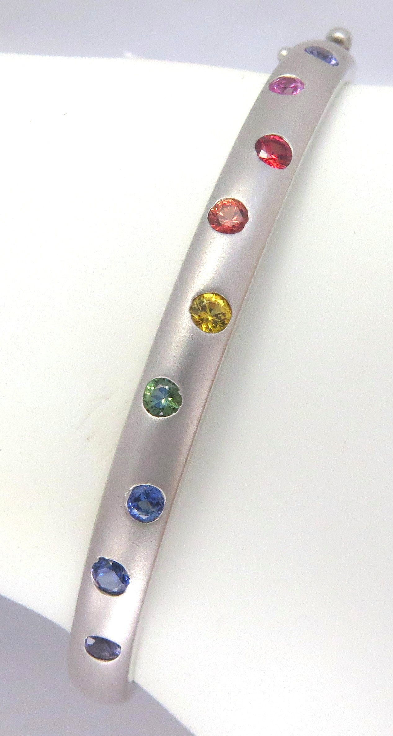 A burnished set rainbow sapphire bangle bracelet in white gold with a brushed finish, displayed on a white background.
