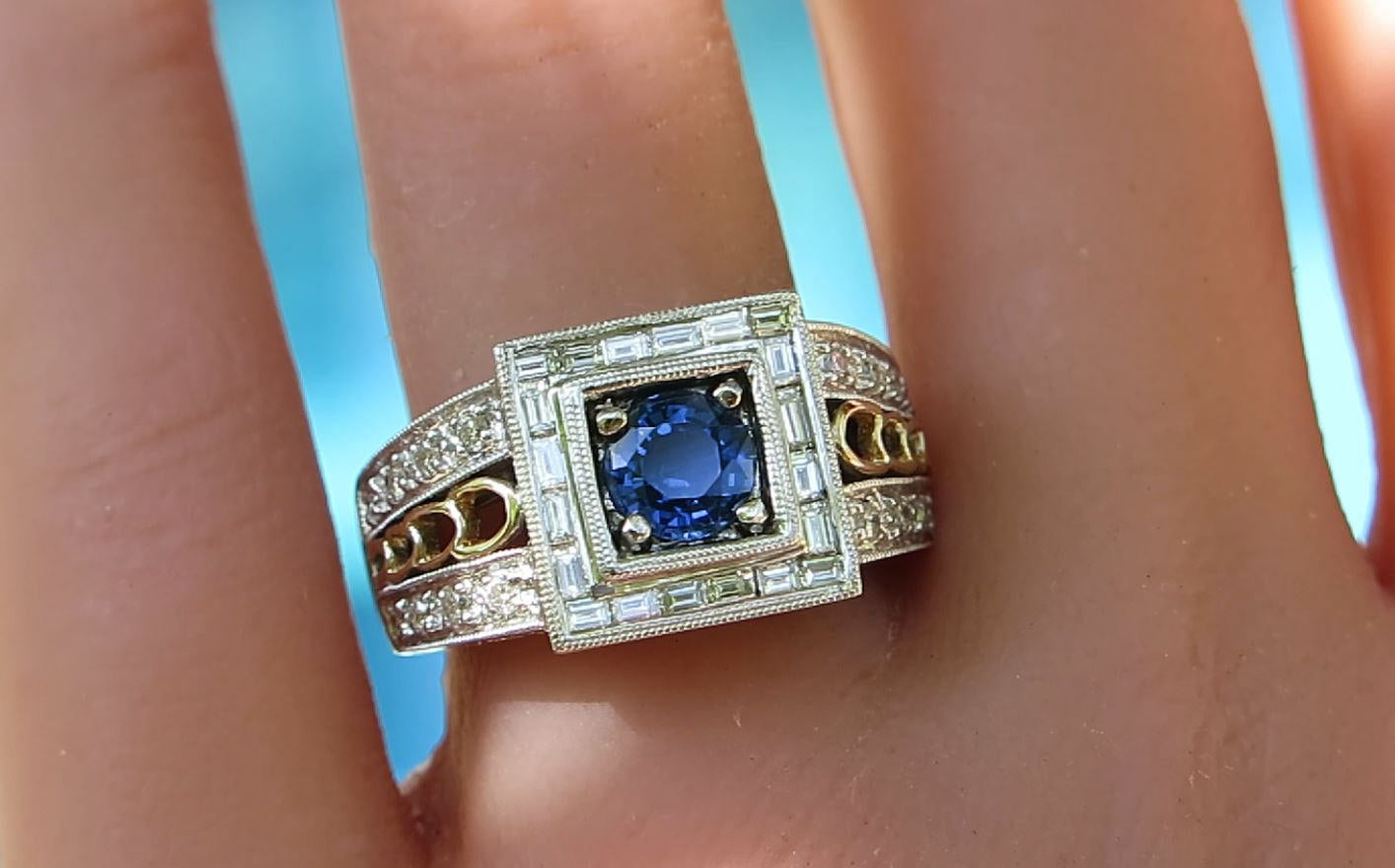 An Art Deco style ring with a round blue sapphire center stone, surrounded by baguette and round diamonds, set on a silver-toned band with gold accents, shown worn on a finger.