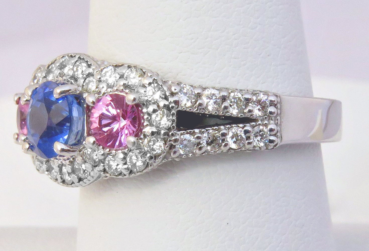 A silver-tone ring with a three-stone cluster featuring a blue sapphire, a pink tourmaline, and clear round diamonds, displayed on a white ring stand.