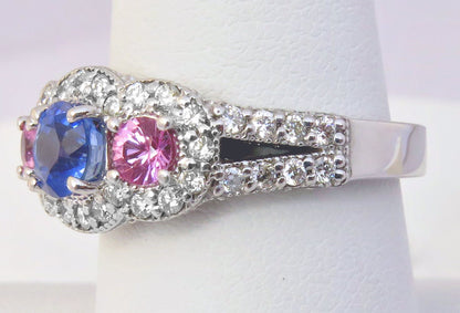 A silver-tone ring with a three-stone cluster featuring a blue sapphire, a pink tourmaline, and clear round diamonds, displayed on a white ring stand.