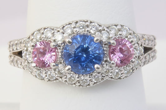 A white gold ring with a central round blue sapphire flanked by two round pink sapphires, all surrounded by a halo of small clear diamonds, displayed on a white ring stand.