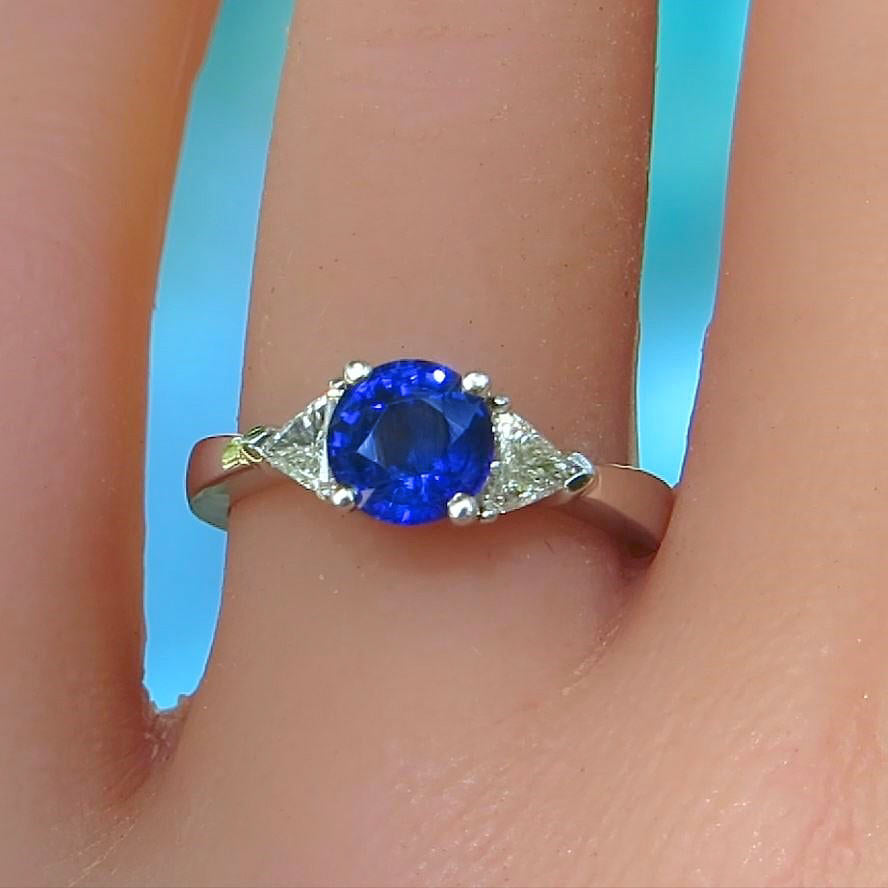 A white gold ring with a round-cut blue sapphire center stone and triangular clear diamond accent stones, shown worn on a finger against a bright blue background.