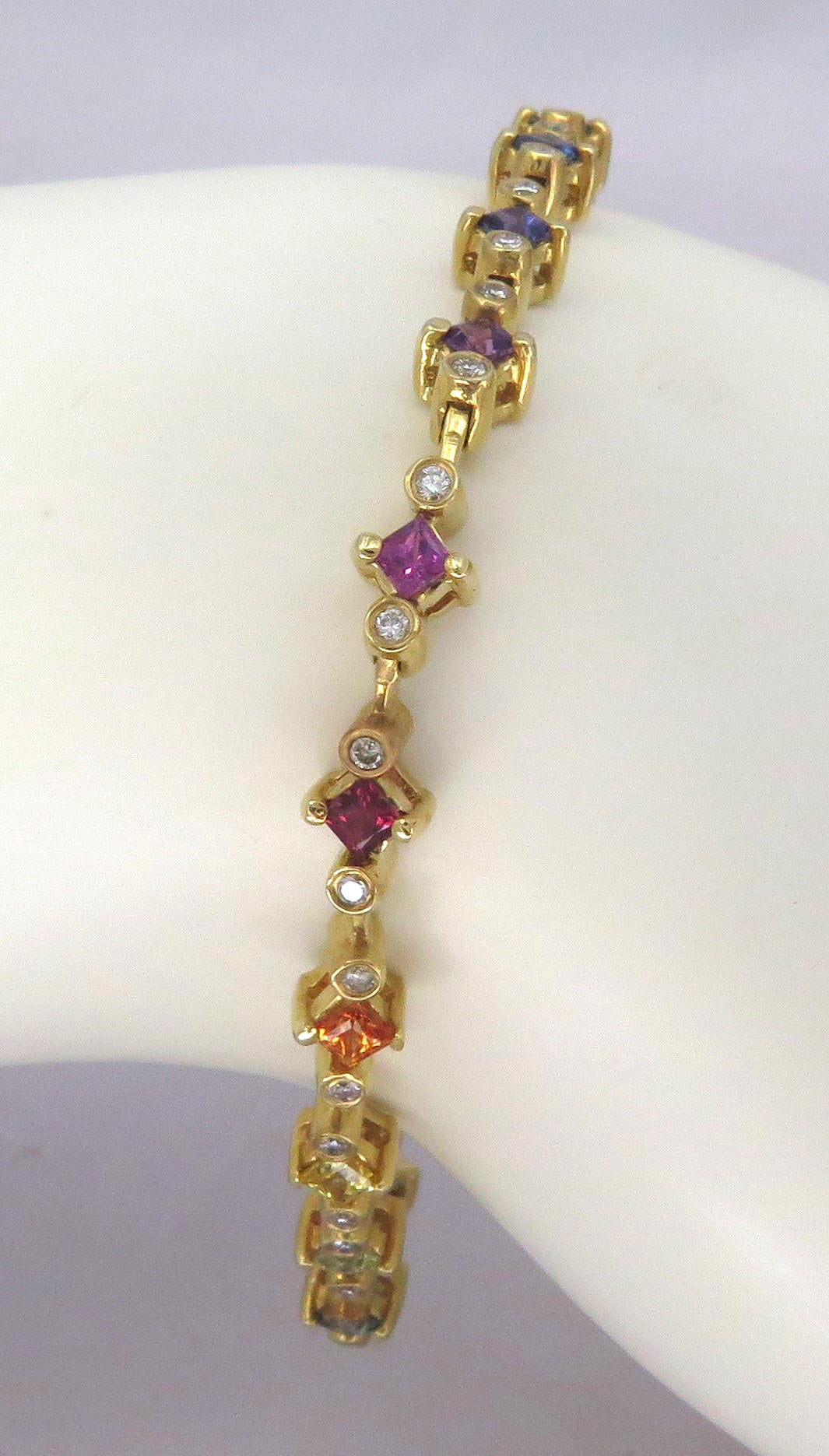 A gold tennis bracelet with alternating colored sapphire gemstone links, displayed on a white mannequin arm. The bracelet features purple, orange, and blue gemstones alongside clear diamonds.