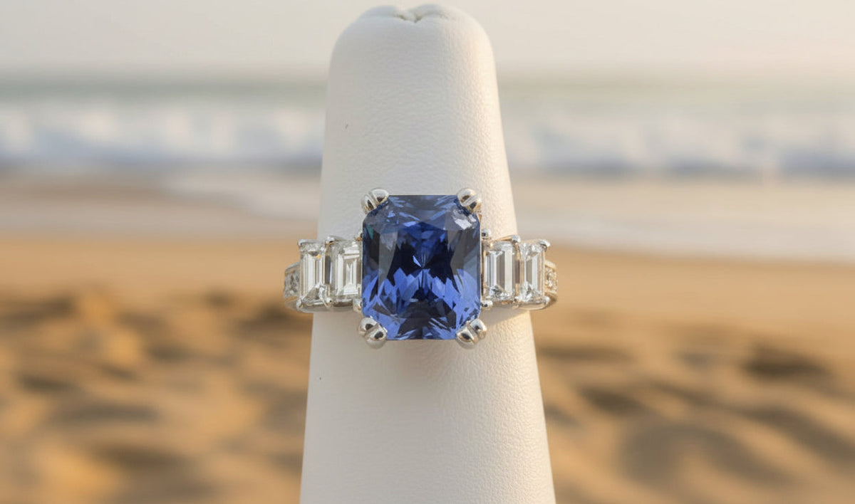 A white gold ring featuring a large rectangular blue sapphire center stone with baguette-cut clear side stones, displayed on a white ring stand.