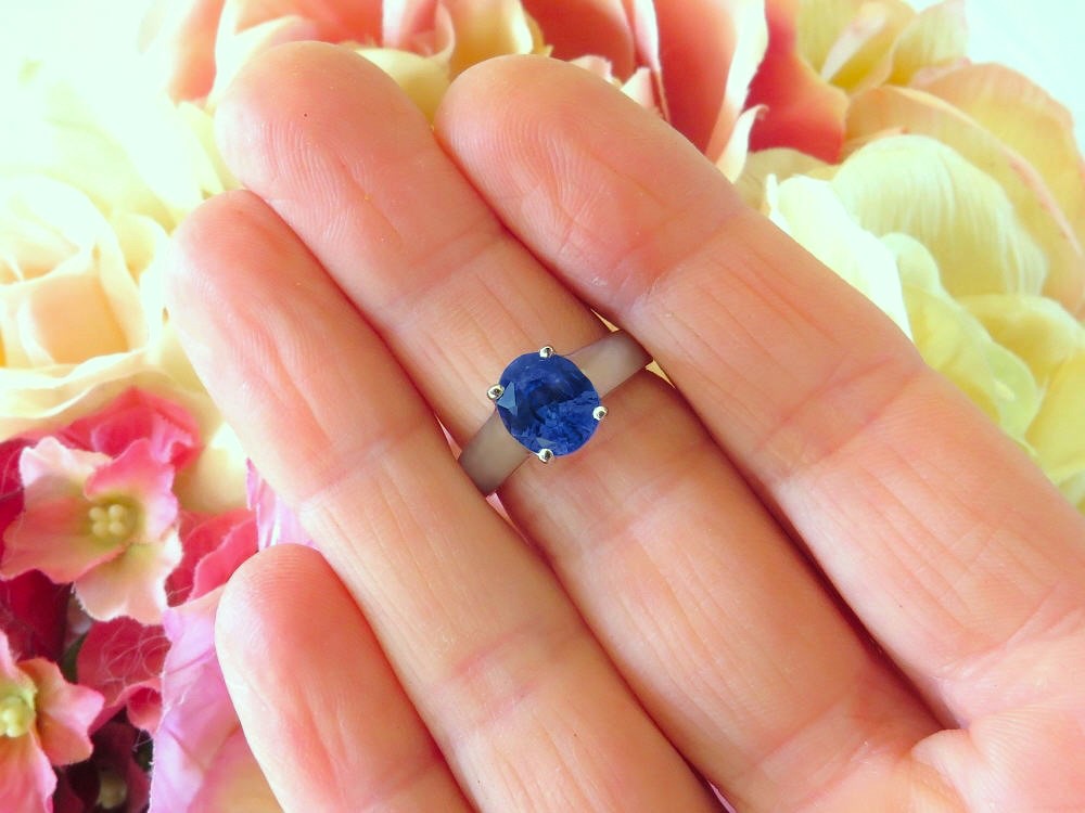 A close-up of a hand wearing a white metal ring with a round-cut deep blue gemstone, set against a soft-focus background of pink and yellow flowers.