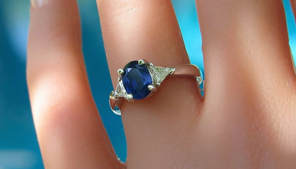 A close-up of a white gold ring with an oval blue sapphire and small side diamonds, worn on a finger against a bright blue background.