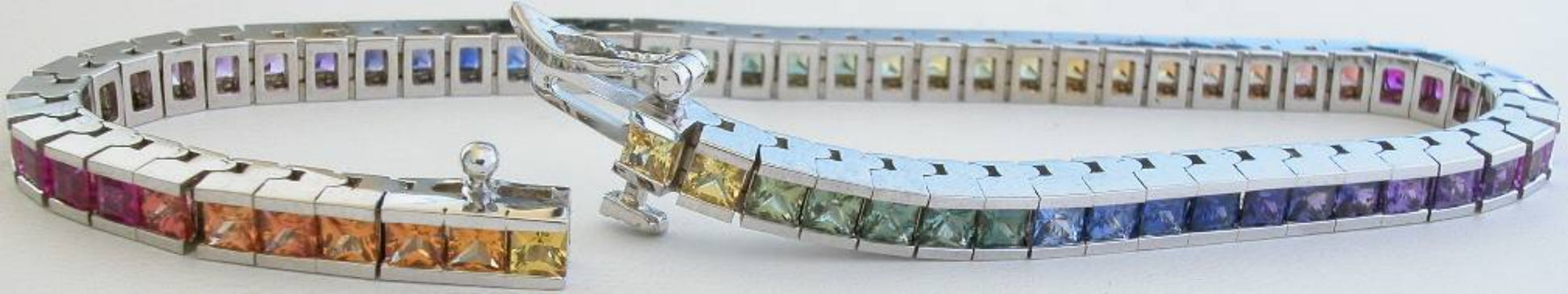 A white gold tennis bracelet with a multi-color sapphire gemstone inlay, shown in a close-up studio shot against a white background.