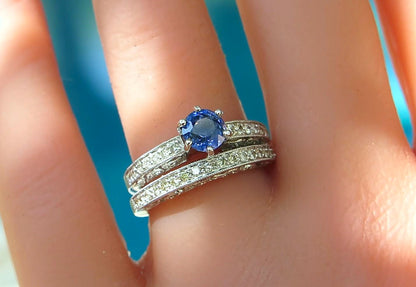 A close-up of a antique style white gold ring and wedding band with a round blue sapphire center stone and small clear stones along the bands, worn on a finger.