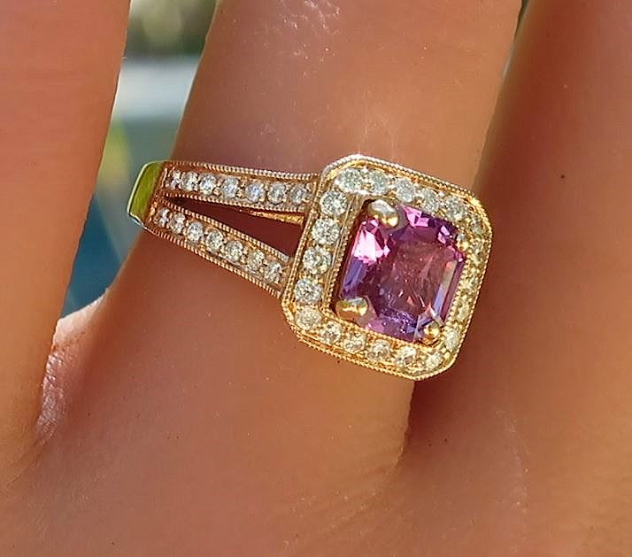 A close-up of a rose gold ring with a square-cut pink magenta sapphire and small clear diamonds surrounding the sapphire, worn on a finger.