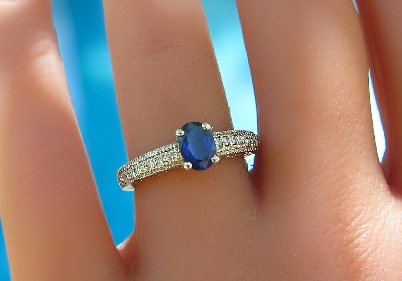 A close-up of a white gold ring with an oval blue sapphire center stone and pavé-set small diamonds, worn on a finger against a bright blue background.