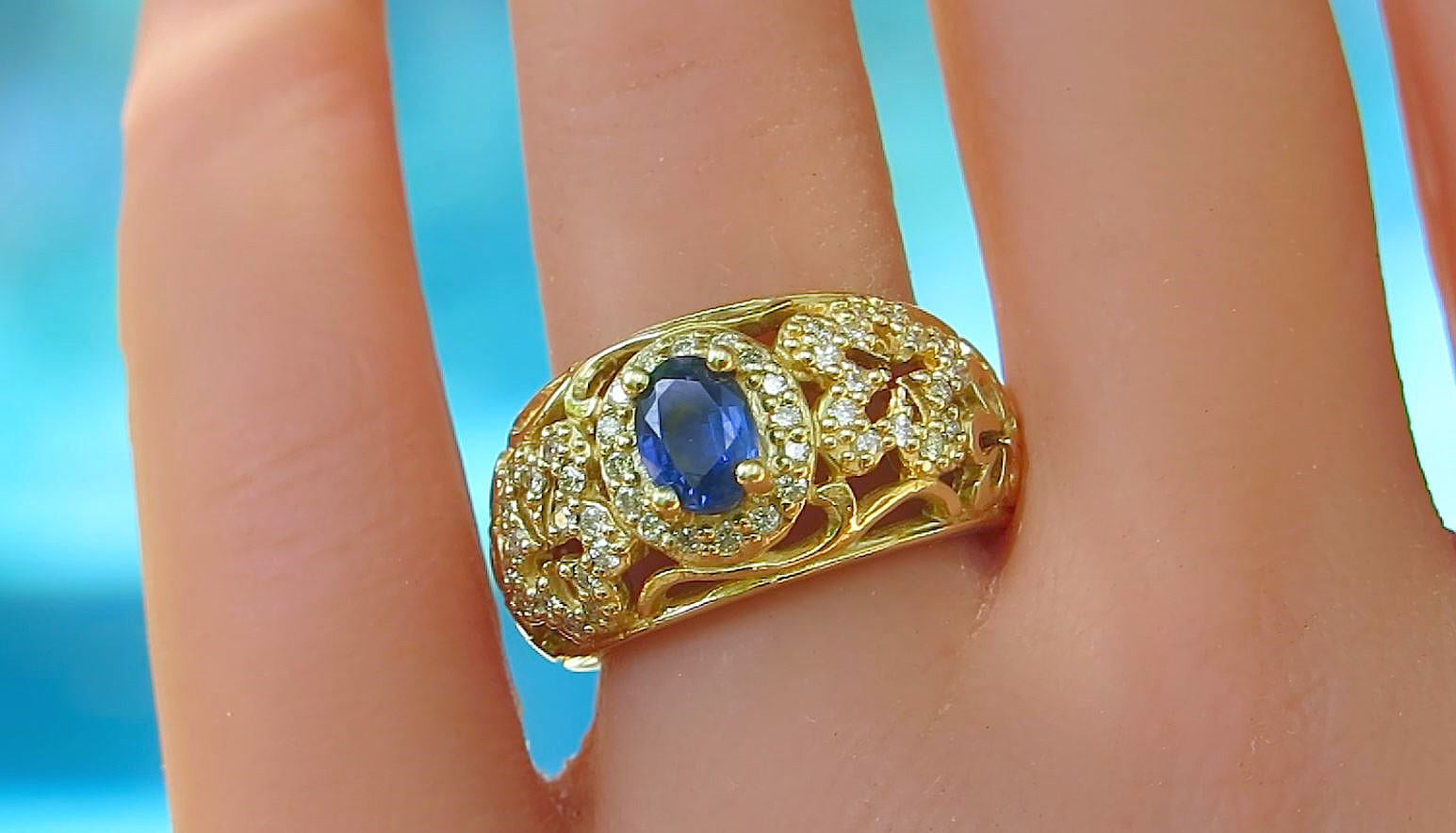 A gold ring with an oval blue sapphire center stone and small clear diamonds set in a flower pattern, shown worn on a finger against a bright blue background.